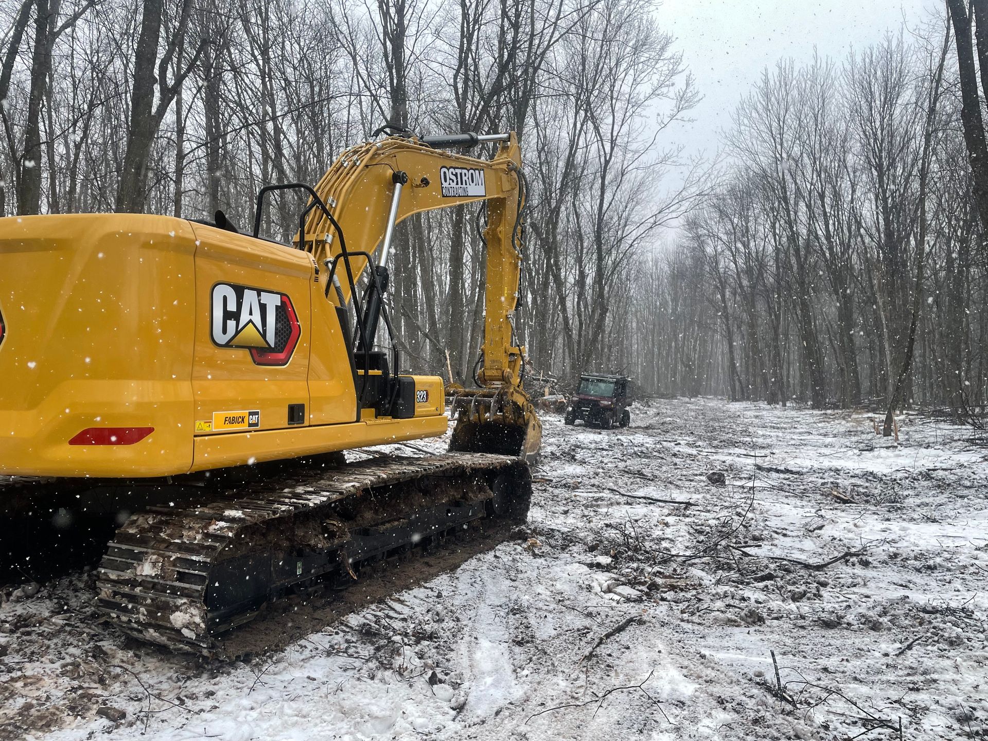A yellow cat excavator is parked on a snowy road in the woods.