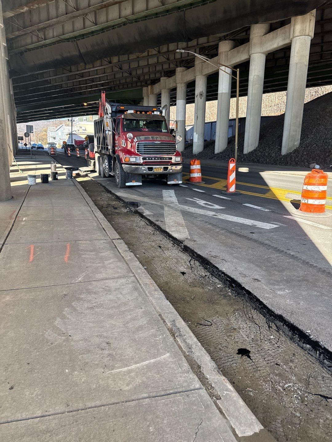 A construction truck under a bridge repairs road asphalt next to a sidewalk with orange cones.