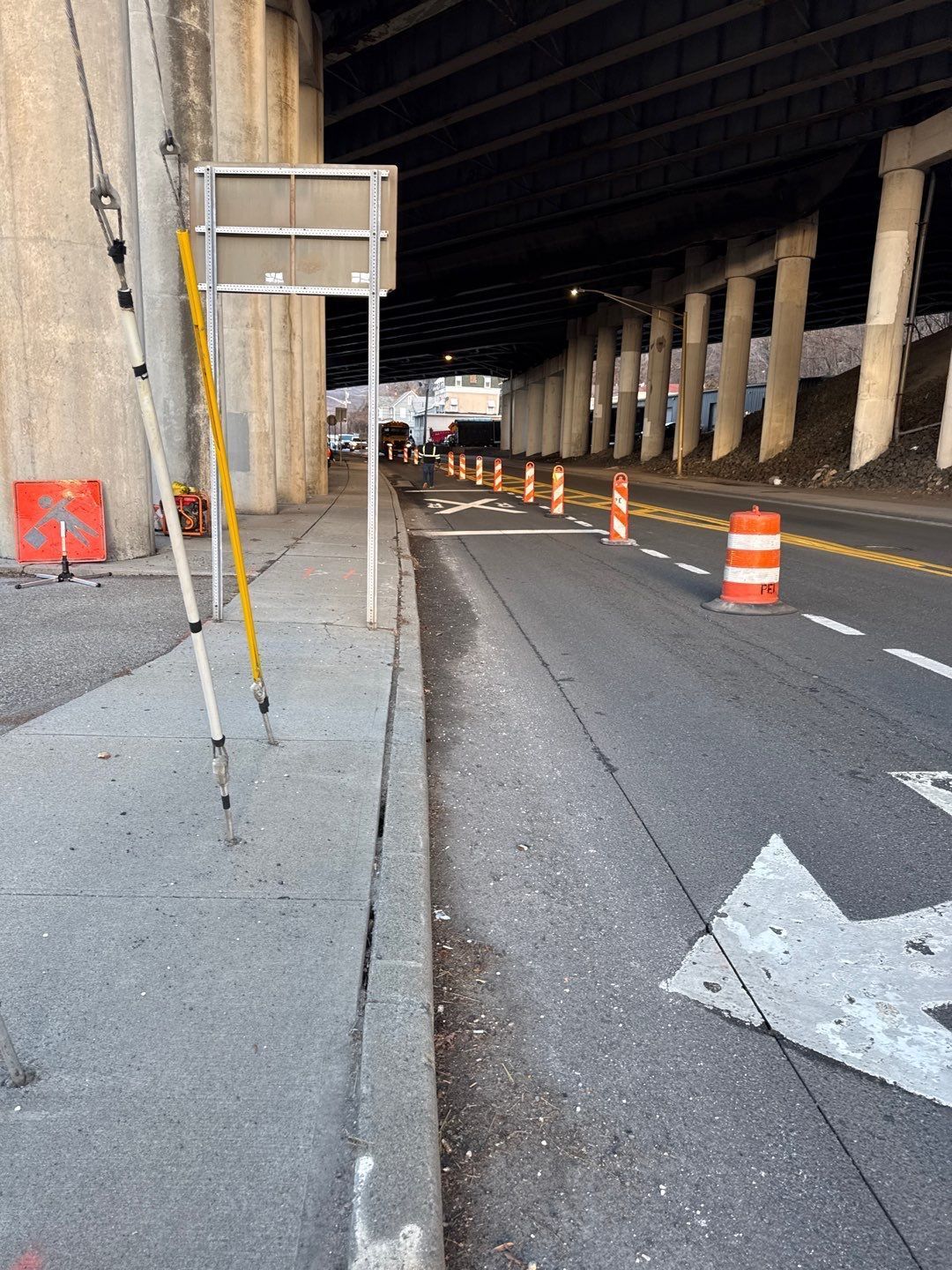 Road under an overpass with construction cones and lane markings. Sidewalk on the left.