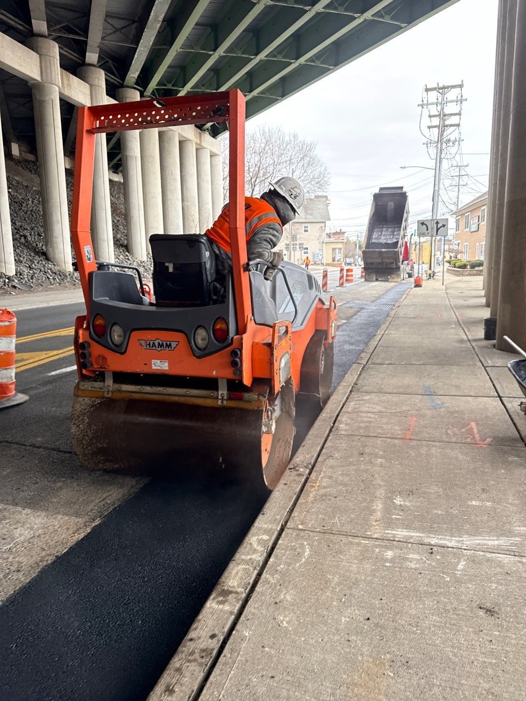 Road worker operating an orange roller compacting fresh asphalt on a road under an overpass.