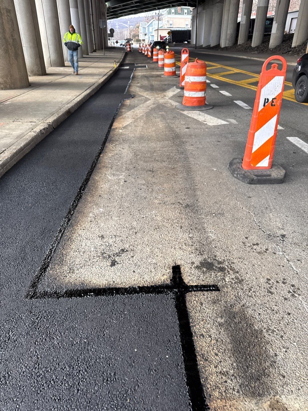 Road construction under an overpass fresh asphalt, orange cones, worker, light traffic.