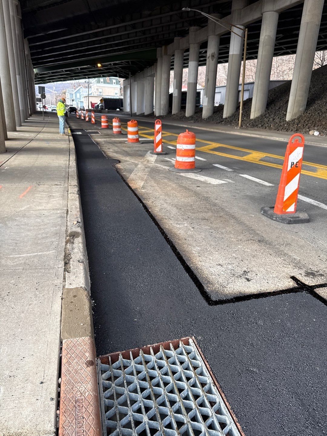 Road construction under an overpass. Newly paved asphalt bordered by concrete with traffic cones. Person in background.