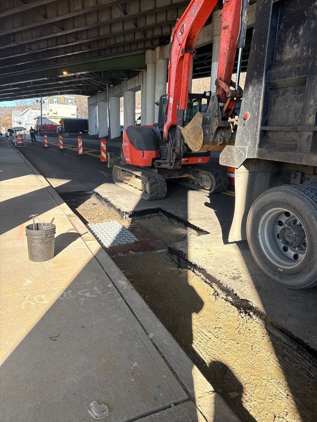 Construction site under a bridge. Excavator working next to a dump truck, cones in the street.