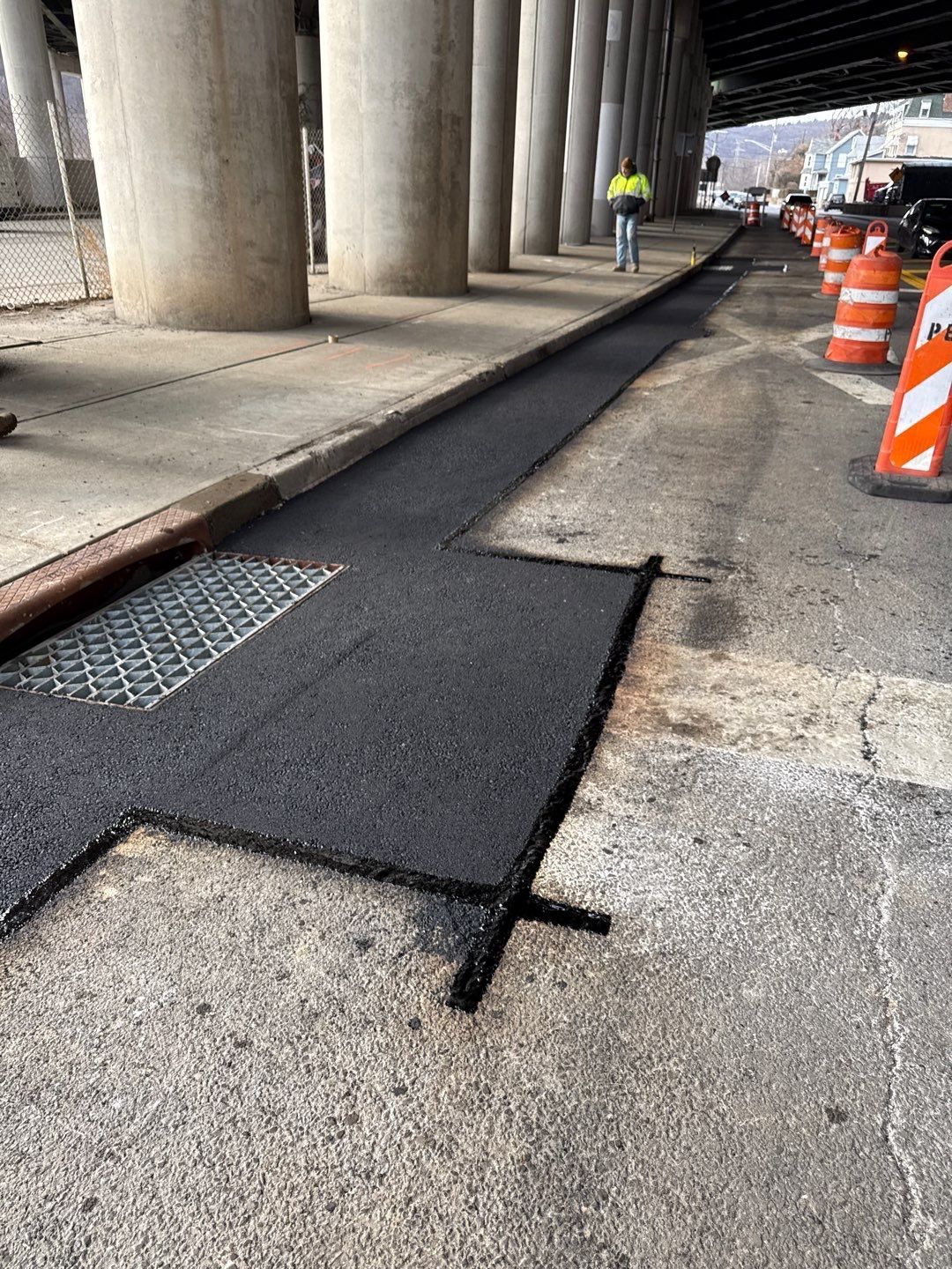 Asphalt paving under a bridge with a worker in the background and traffic cones.