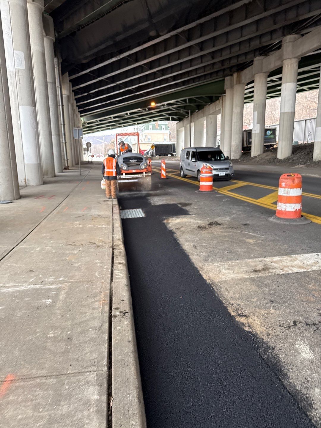 Road work under overpass with workers and vehicles. Fresh asphalt. Orange safety barrels.