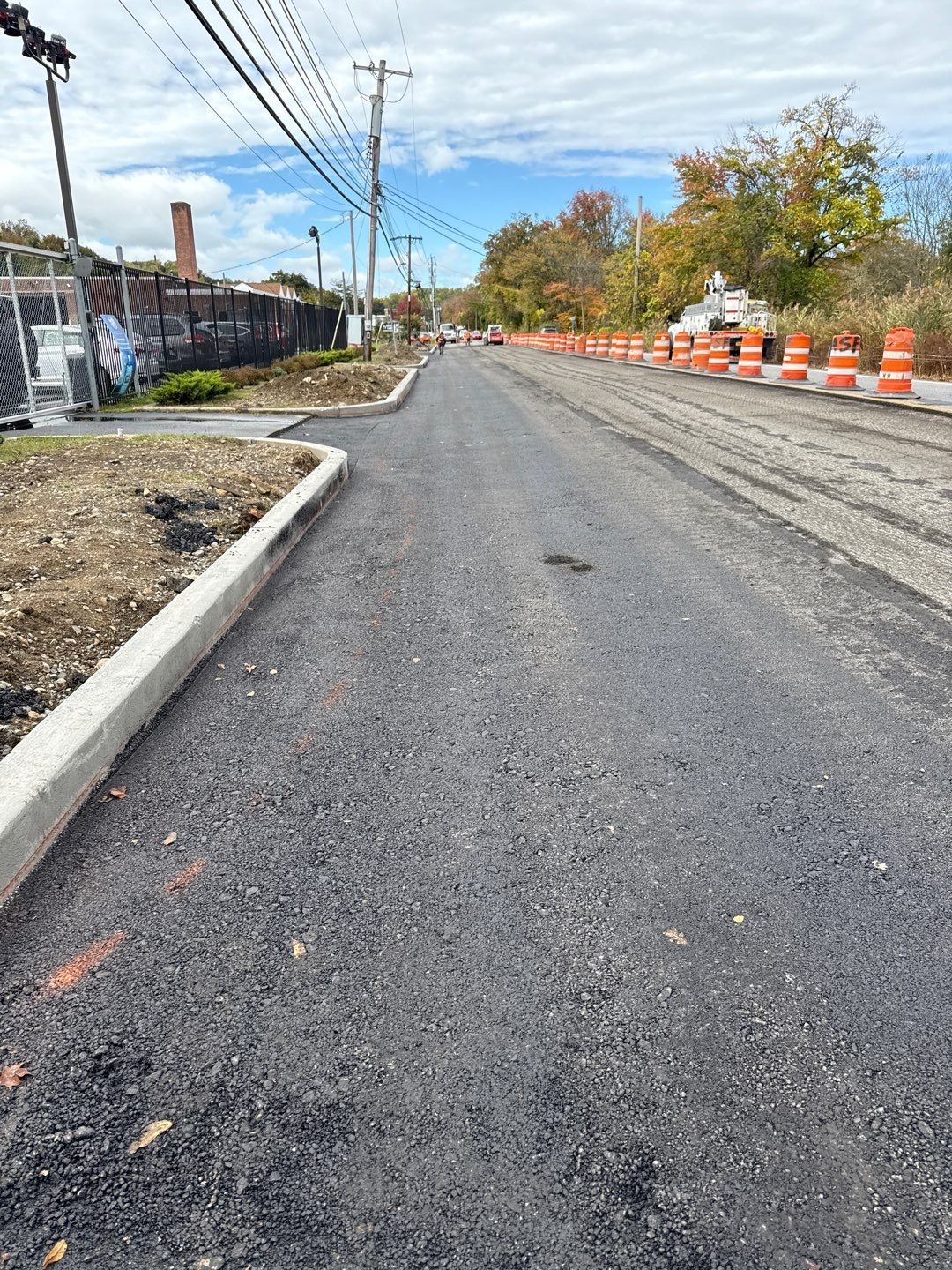 Newly paved road with a concrete curb on the left, orange cones on the right. Blue sky and trees.