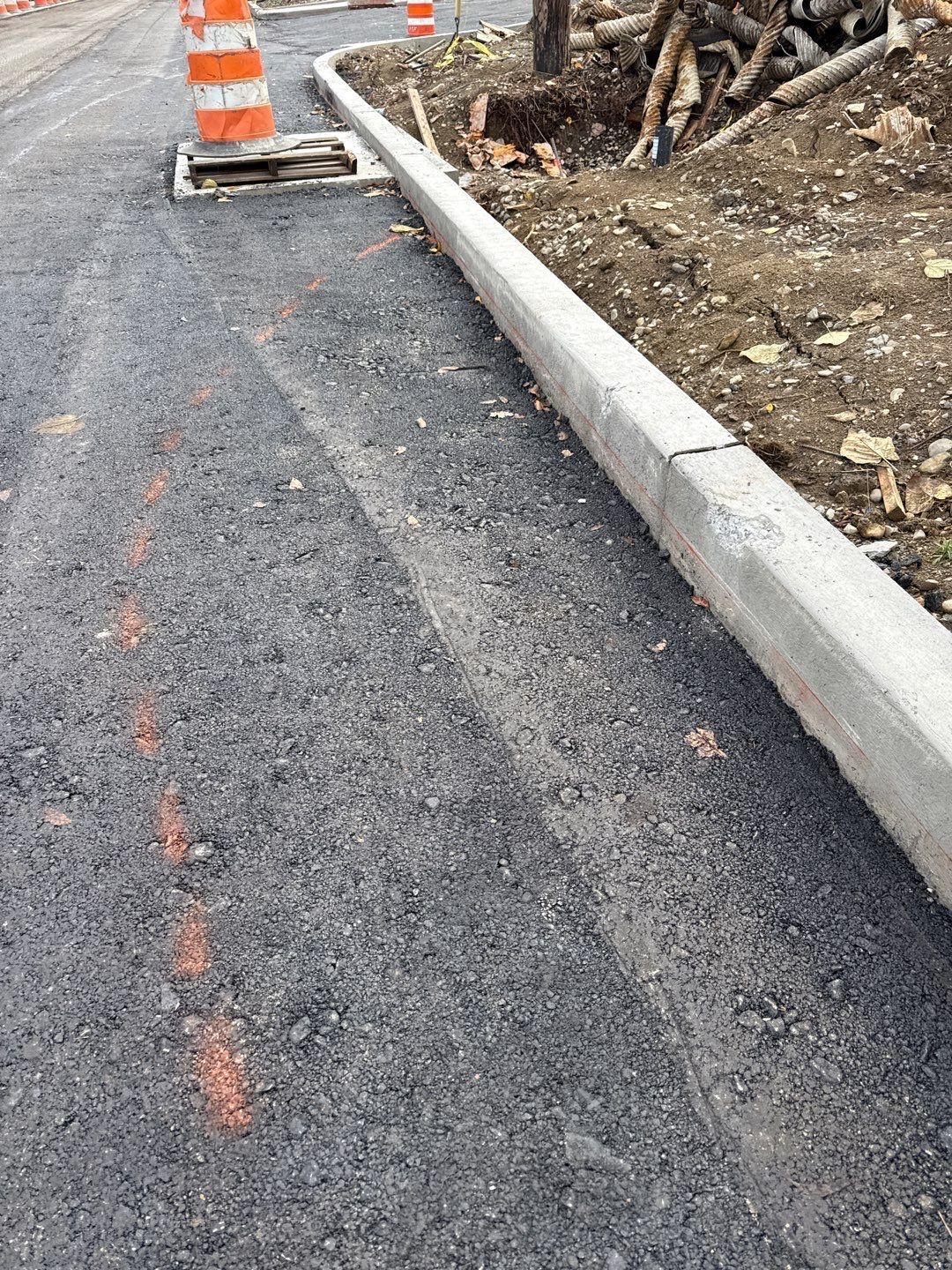 Newly paved asphalt with concrete curb, orange cone, and open utility access in road construction area.