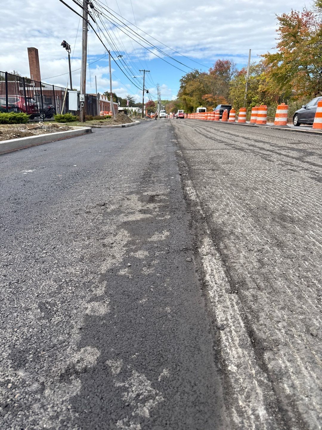 Road undergoing construction; asphalt and worn pavement visible with traffic cones and building in background.