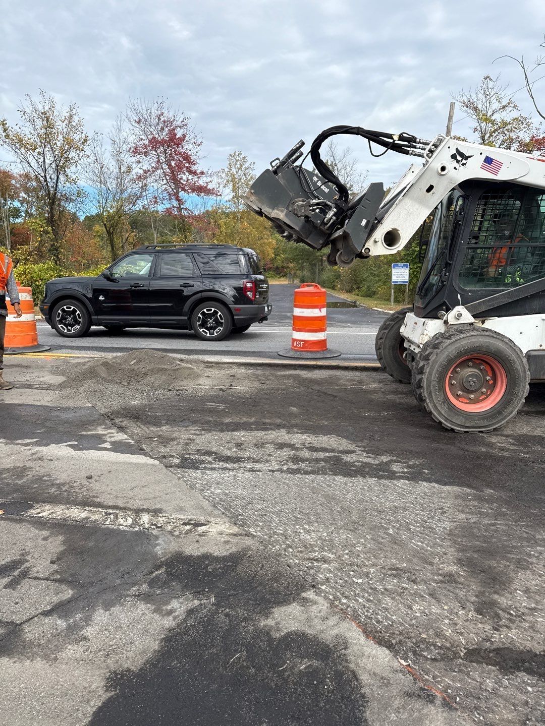 Black SUV near a Bobcat skid-steer loader on a paved road. Orange traffic cones. Trees in the background.