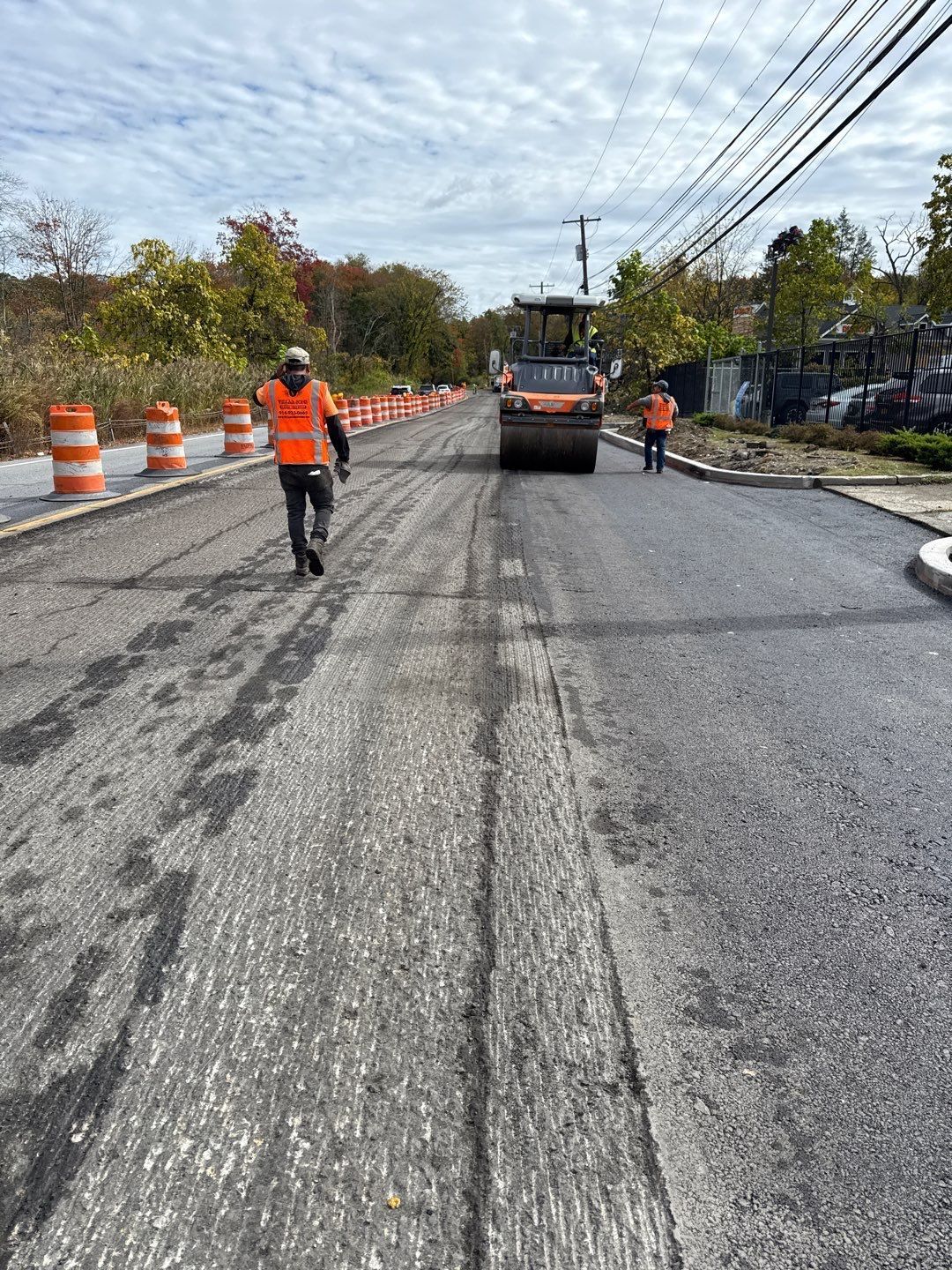Road construction: workers in orange vests, roller compacting asphalt. Barricades line the road under a cloudy sky.