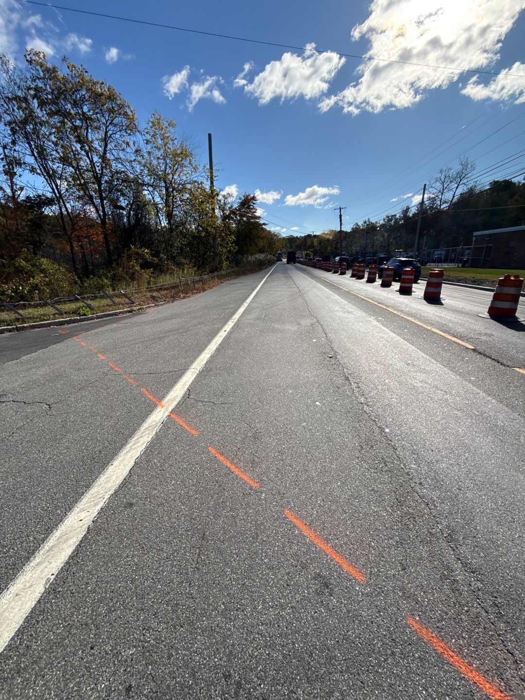 Road under construction; asphalt, orange lines, traffic barrels, and blue sky.