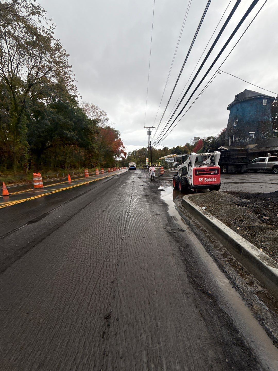 Road construction: freshly paved road, Bobcat, power lines, cloudy sky, orange cones, and a building on the right.