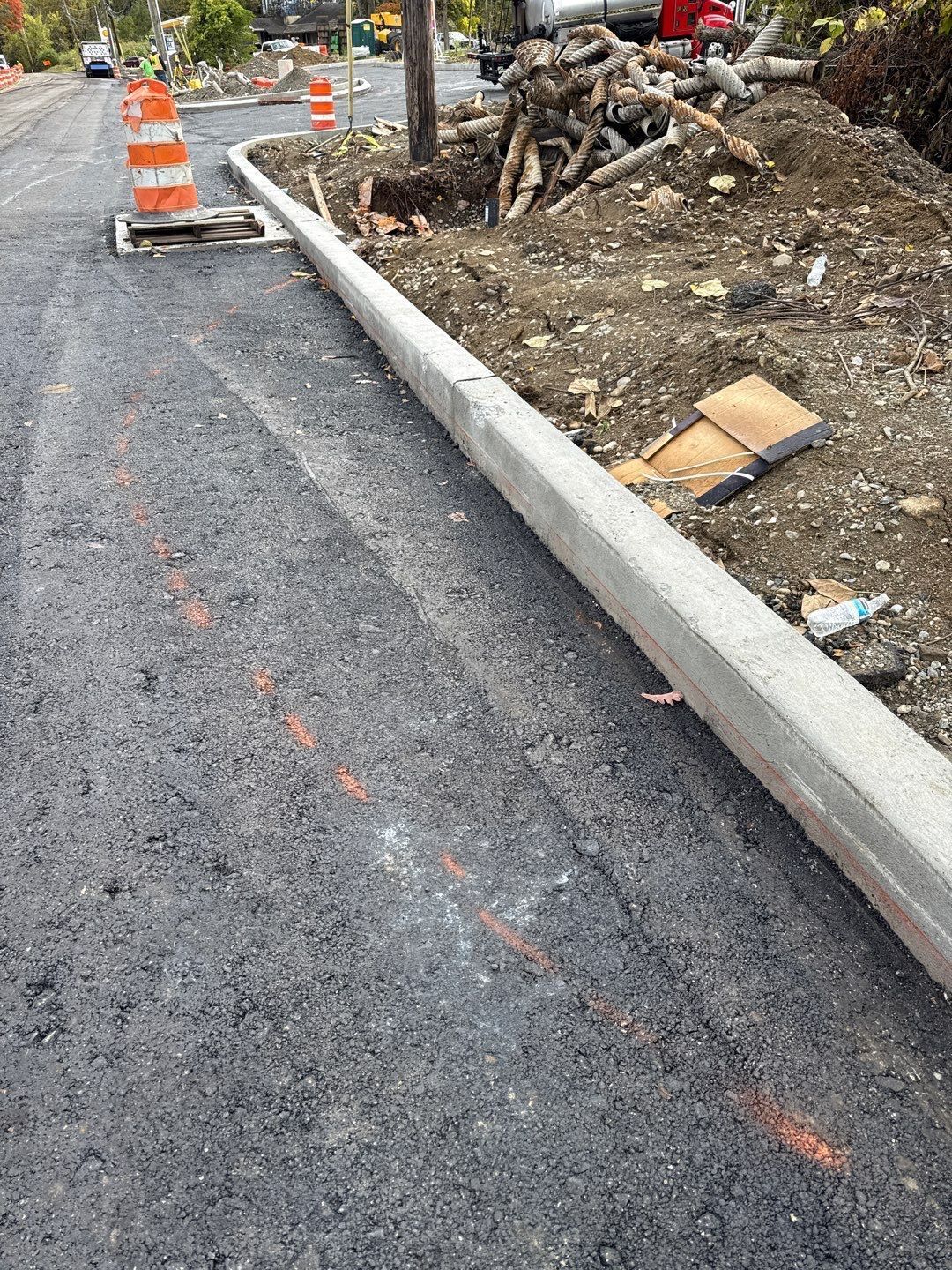Road construction: Asphalt surface, concrete curb, orange safety cone, construction materials on the side.
