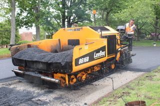 A man is standing next to a yellow and black asphalt paving machine.