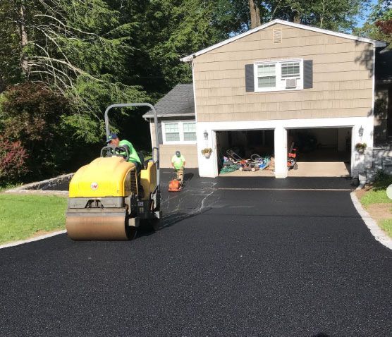 A yellow roller is working on a driveway in front of a house.