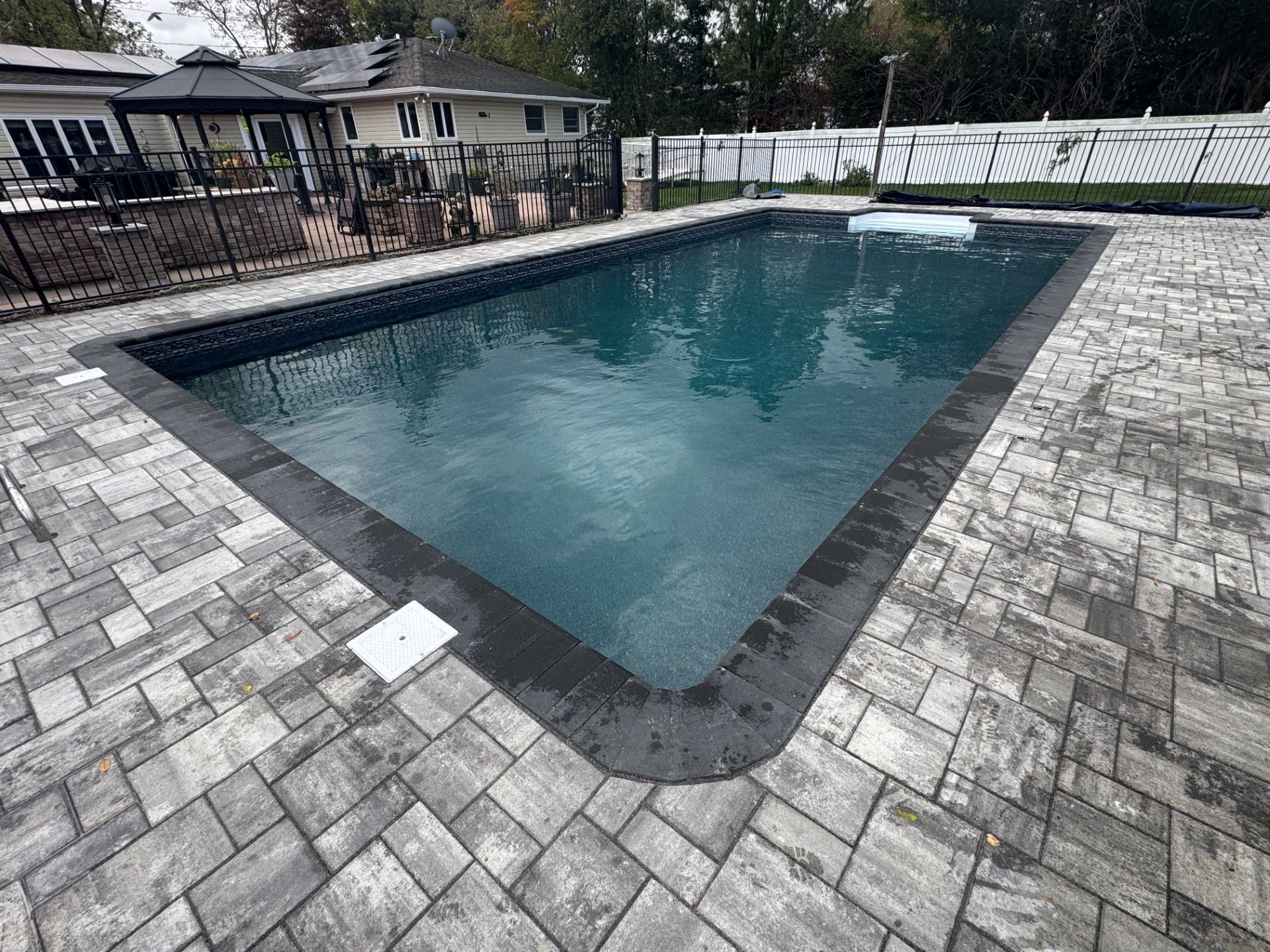 Rectangular pool with dark trim surrounded by gray pavers. A gazebo and house are in the background.