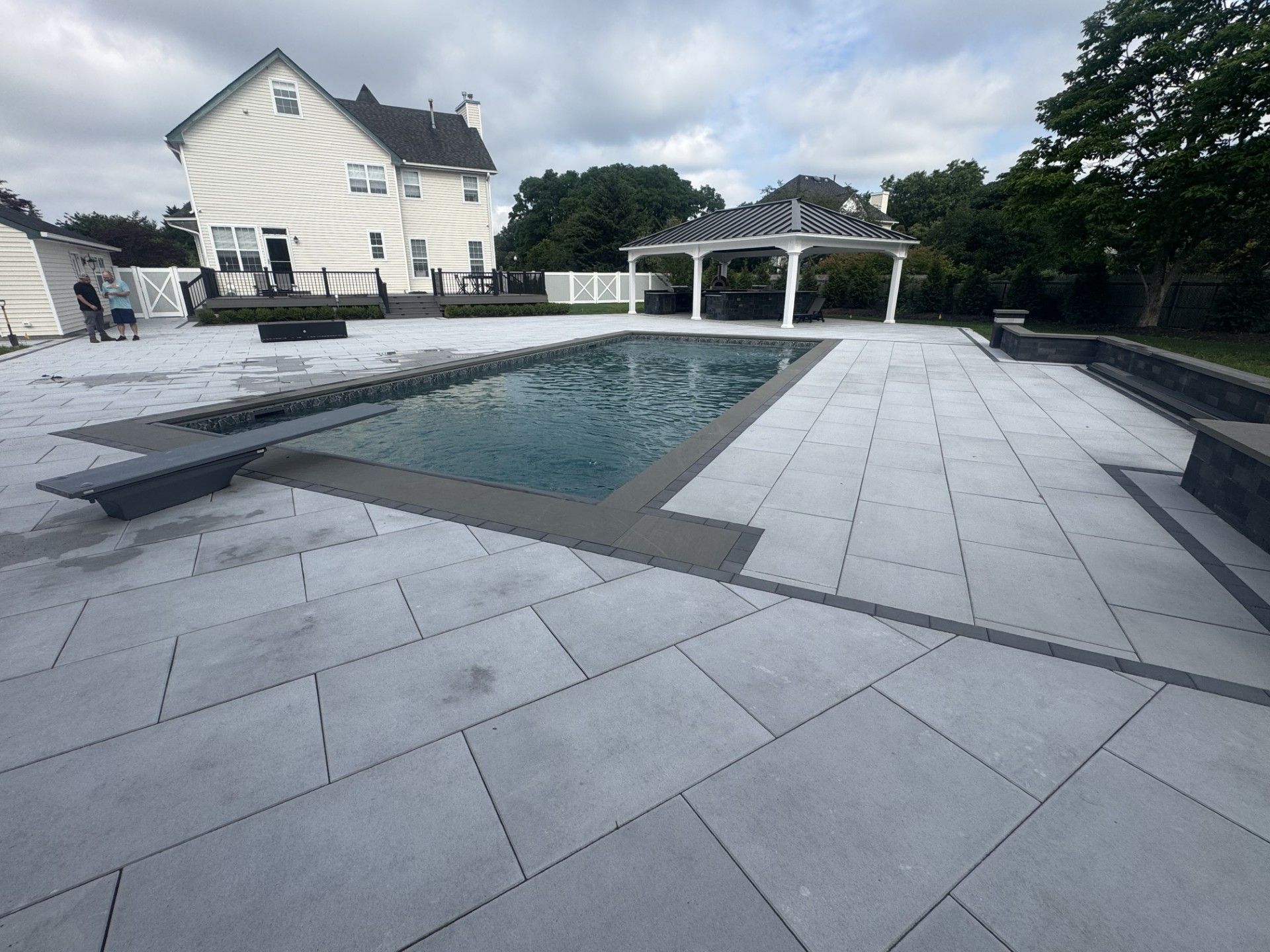 Backyard with pool, gray stone patio, white gazebo, two-story house, and overcast sky.