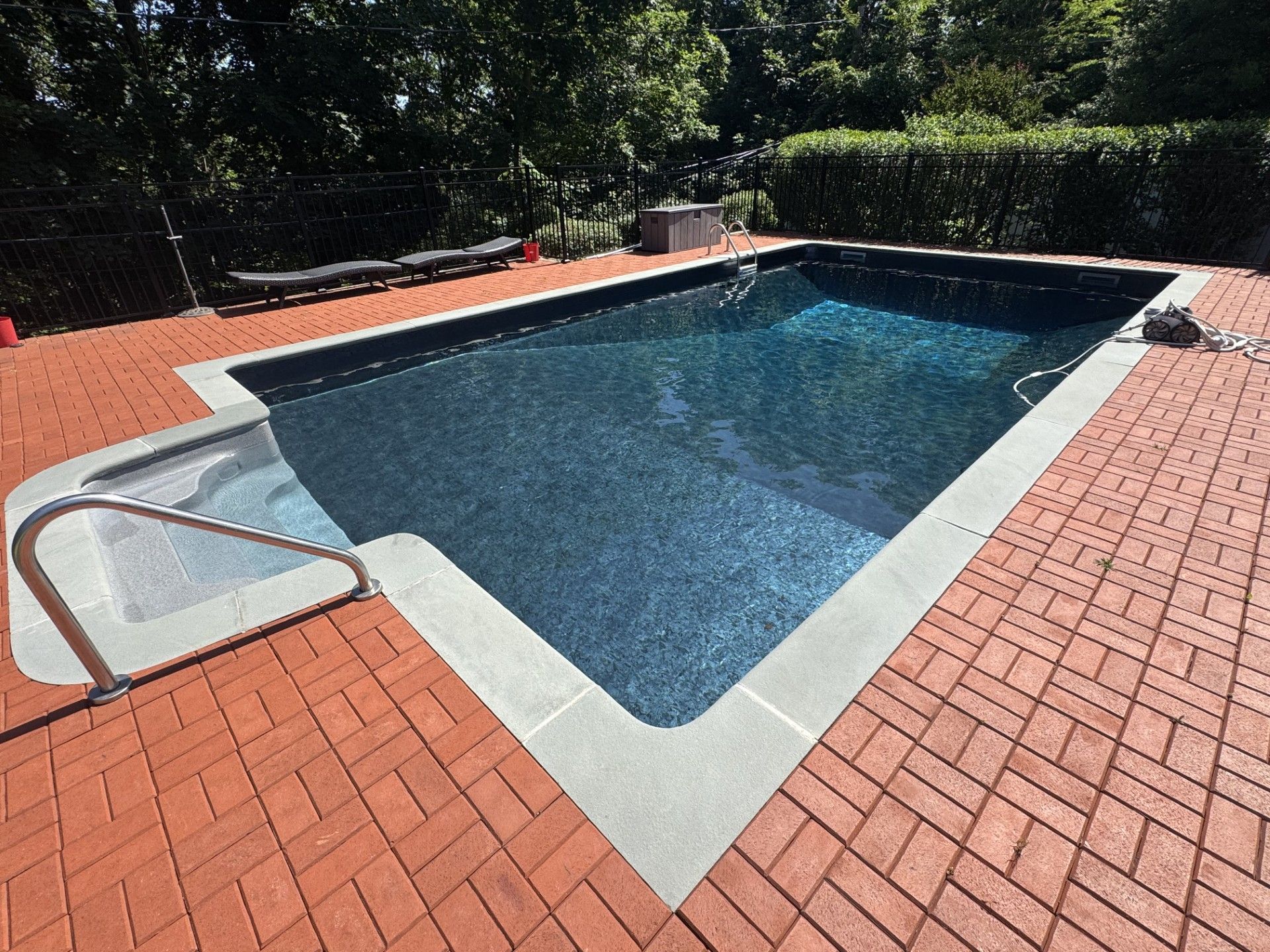 Rectangular pool with attached spa, surrounded by brick patio. Blue water reflects sunlight, trees in the background.