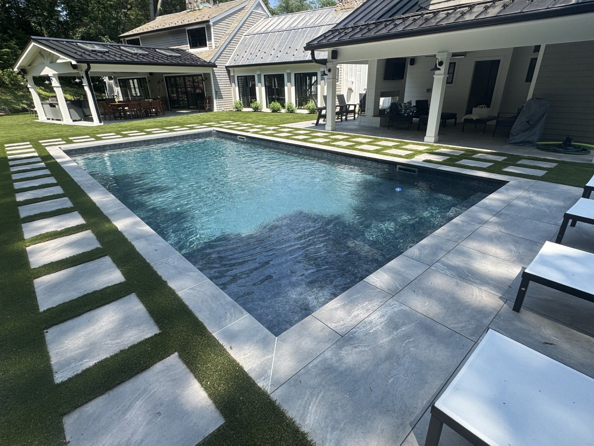 Rectangular pool surrounded by concrete pavers and grass, with a house and patio visible in the background.