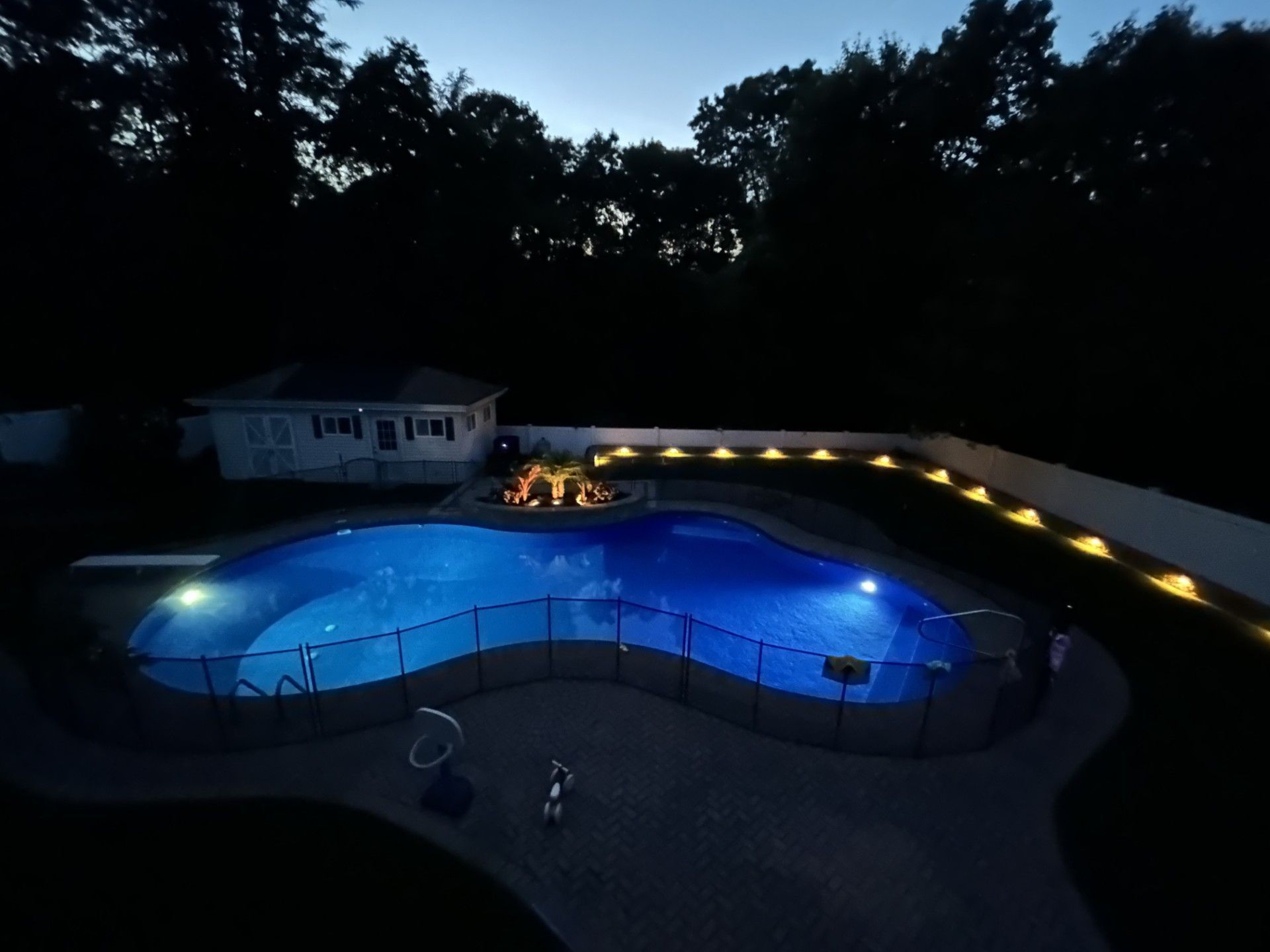 Nighttime view of a backyard pool, illuminated blue. A fire pit glows nearby, pathway lights, and a small house in the background.