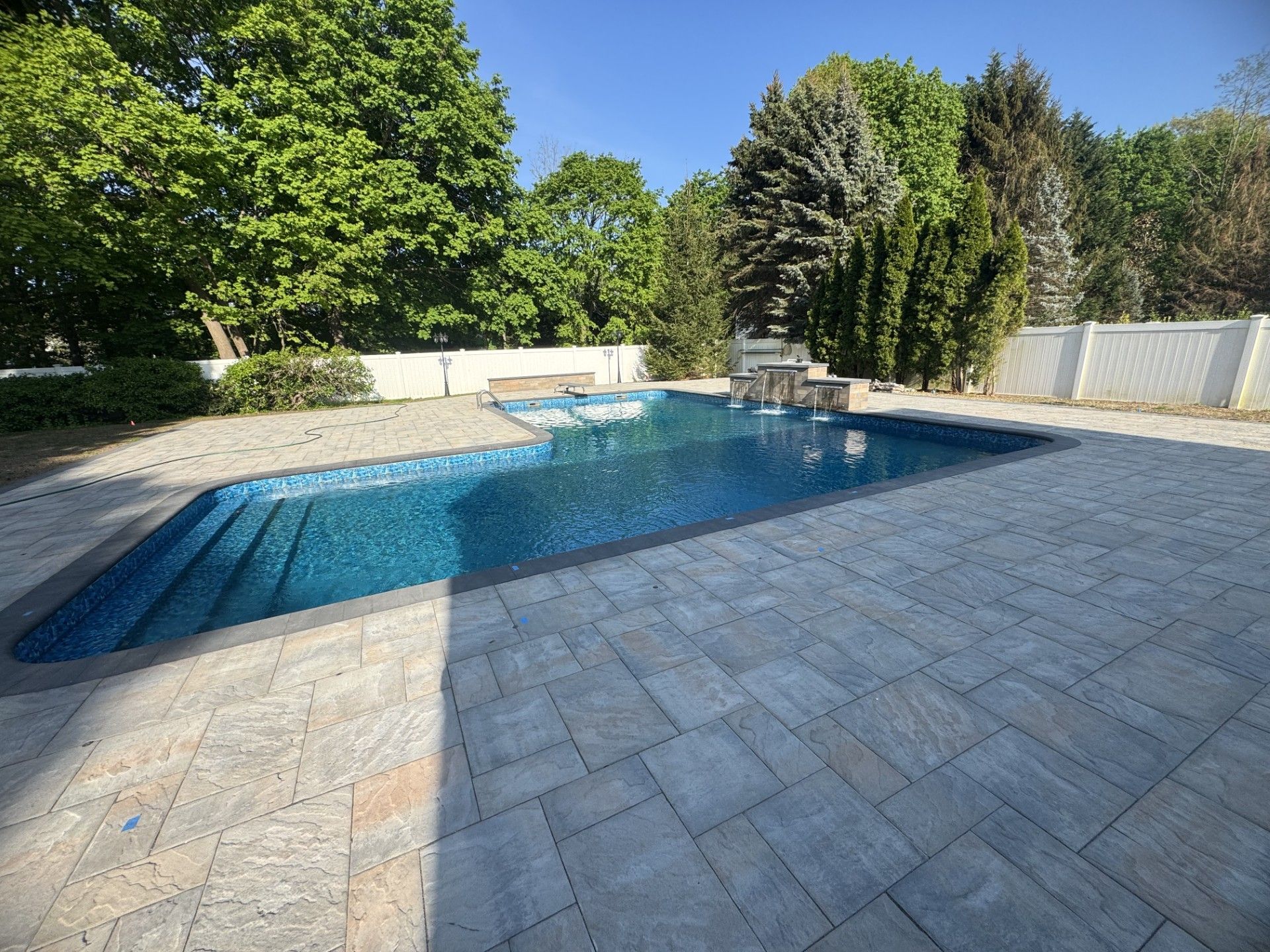 Swimming pool with blue water and stone patio, surrounded by trees and a white fence.