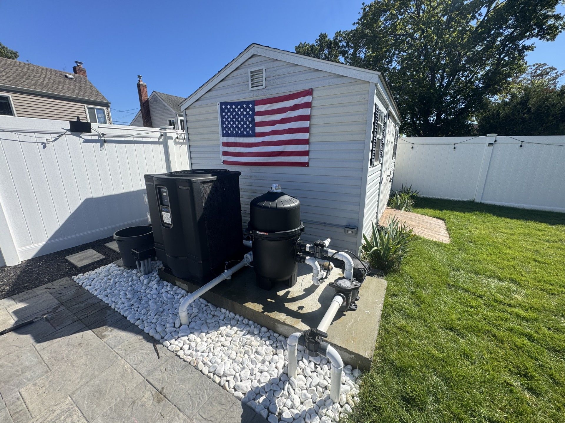 Pool equipment next to a shed with an American flag; a white fence and a lawn surround the area.