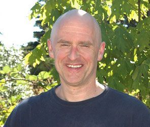 Smiling bald man in a dark shirt standing outdoors in front of leafy green trees
