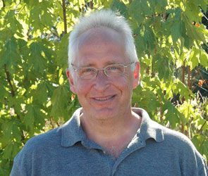 Smiling man with glasses standing outdoors in front of green leaves and sunlight