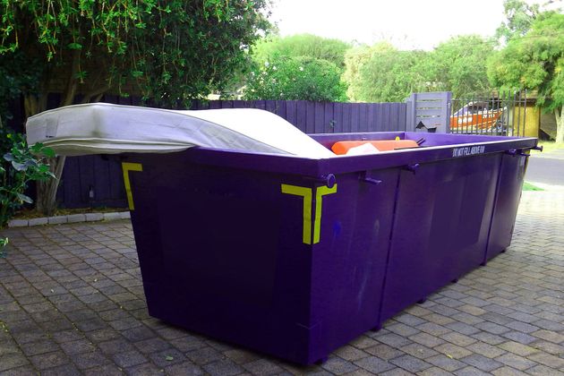 Large purple dumpster on a paved driveway beside a wooden fence and trees
