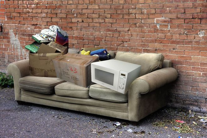 Discarded couch with boxes and a microwave dumped beside a brick wall on a street.
