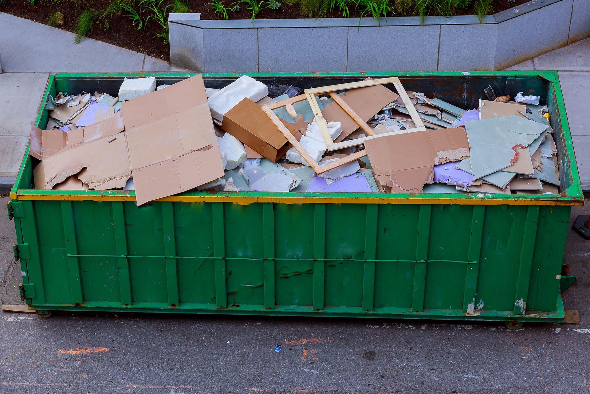 Green dumpster overflowing with cardboard and construction debris on a sidewalk