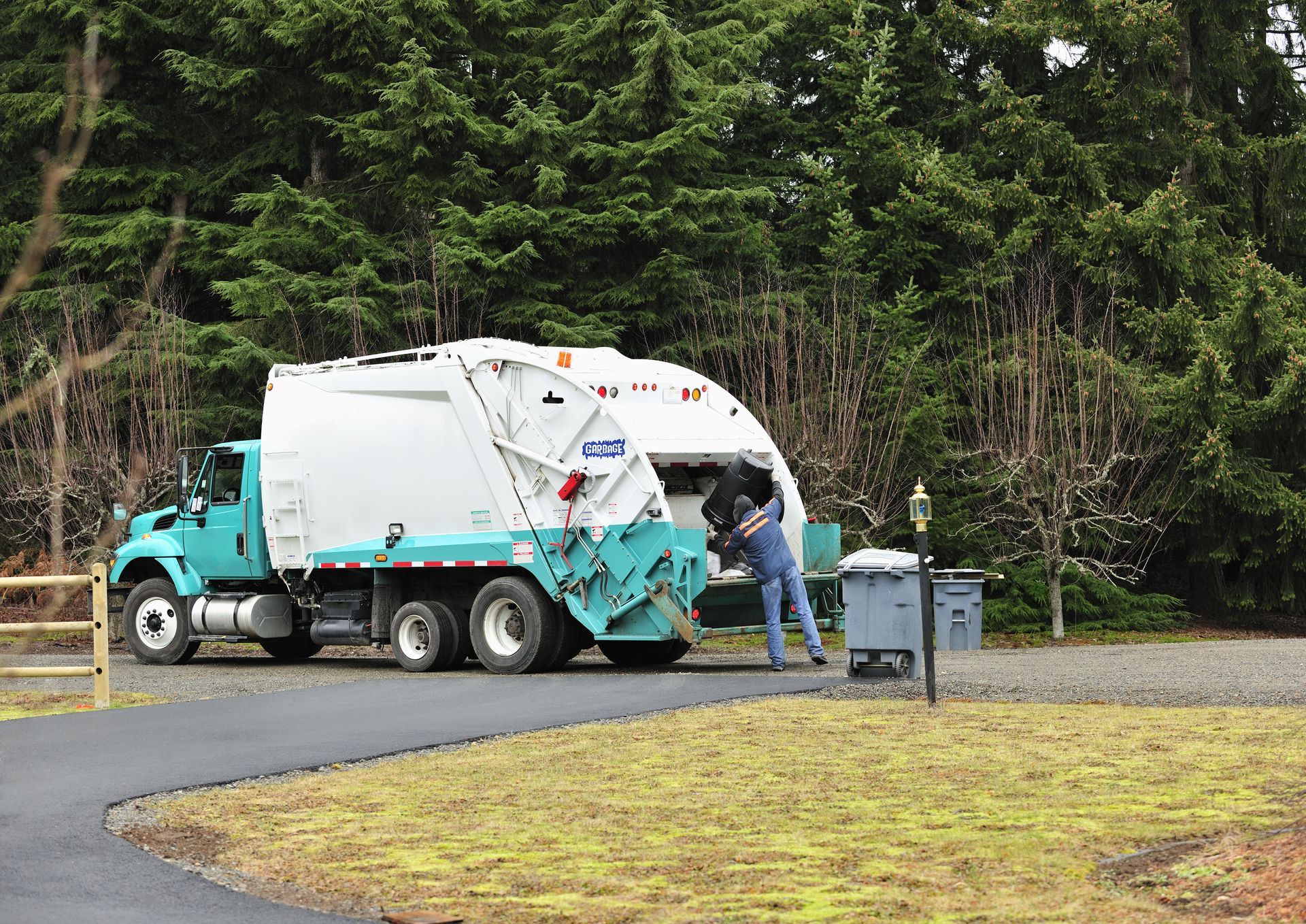 Garbage truck and worker at a roadside bin near a wooded area