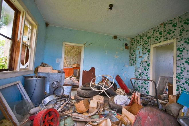 Cluttered, dilapidated room with debris, broken furniture, peeling paint, and a window letting in daylight