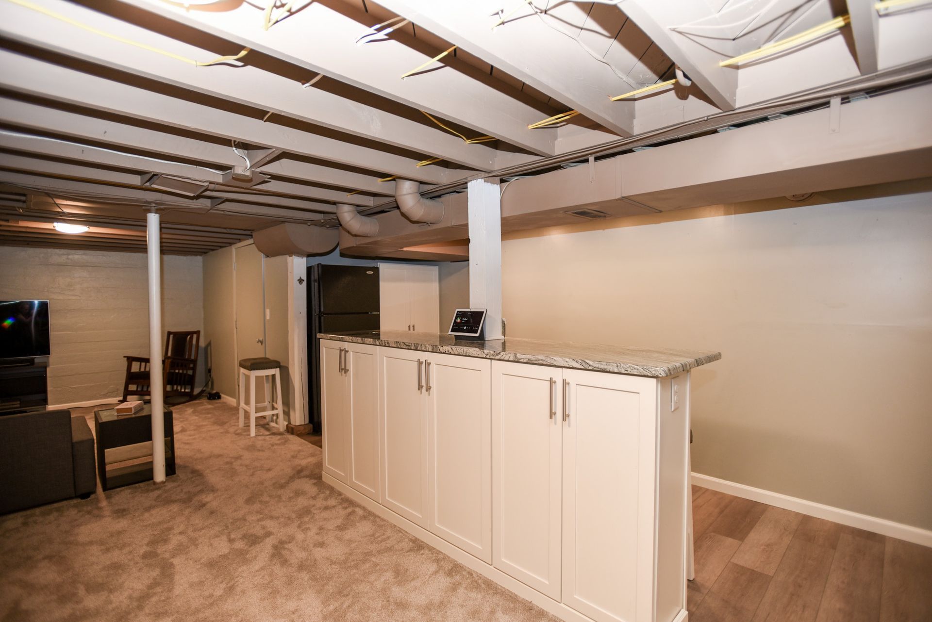 Finished basement with white bar cabinets, exposed ceiling beams, and carpeted flooring.