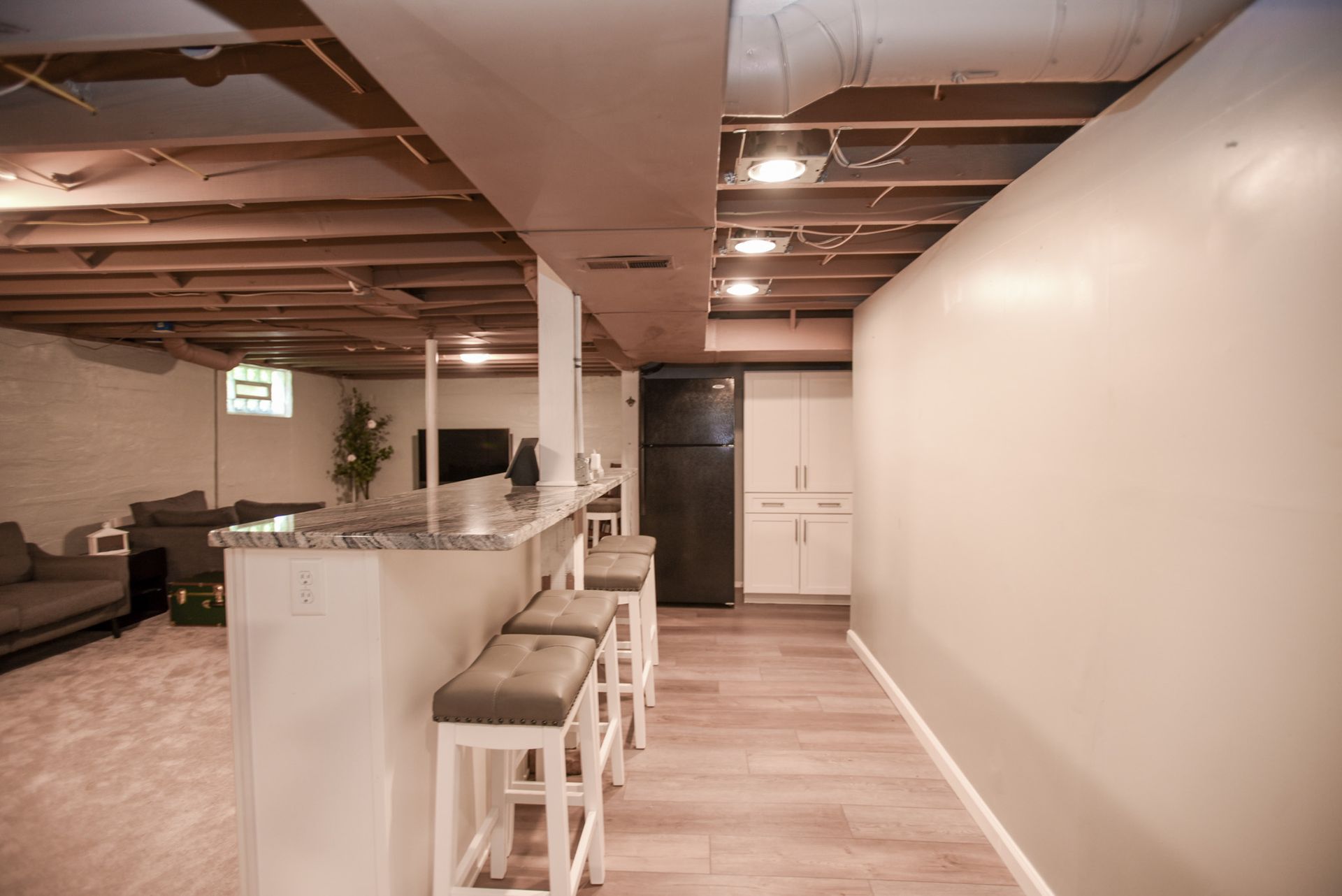 Open-concept basement kitchen and living area with bar stools, white walls, and exposed ceiling beams.