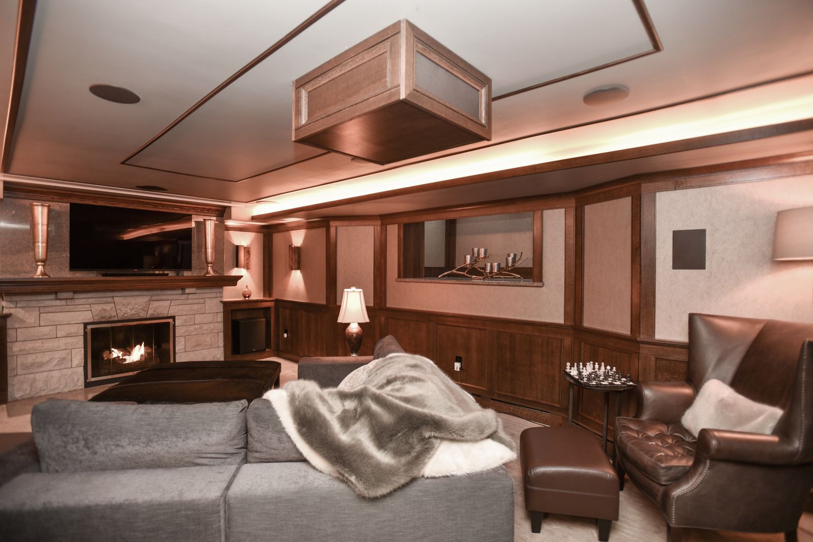Cozy living room with stone fireplace, gray sofa, leather chair, wood paneling, and warm ceiling lighting.