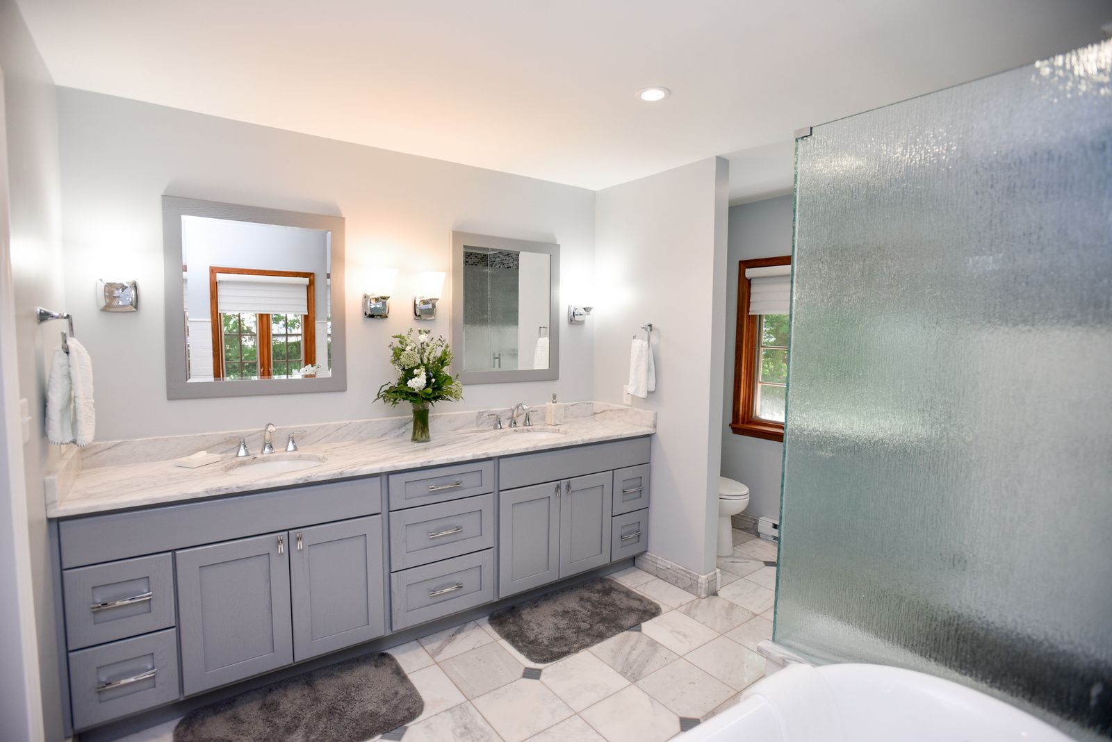 Modern bathroom with gray vanity, marble countertop, mirrors, and frosted glass shower door.