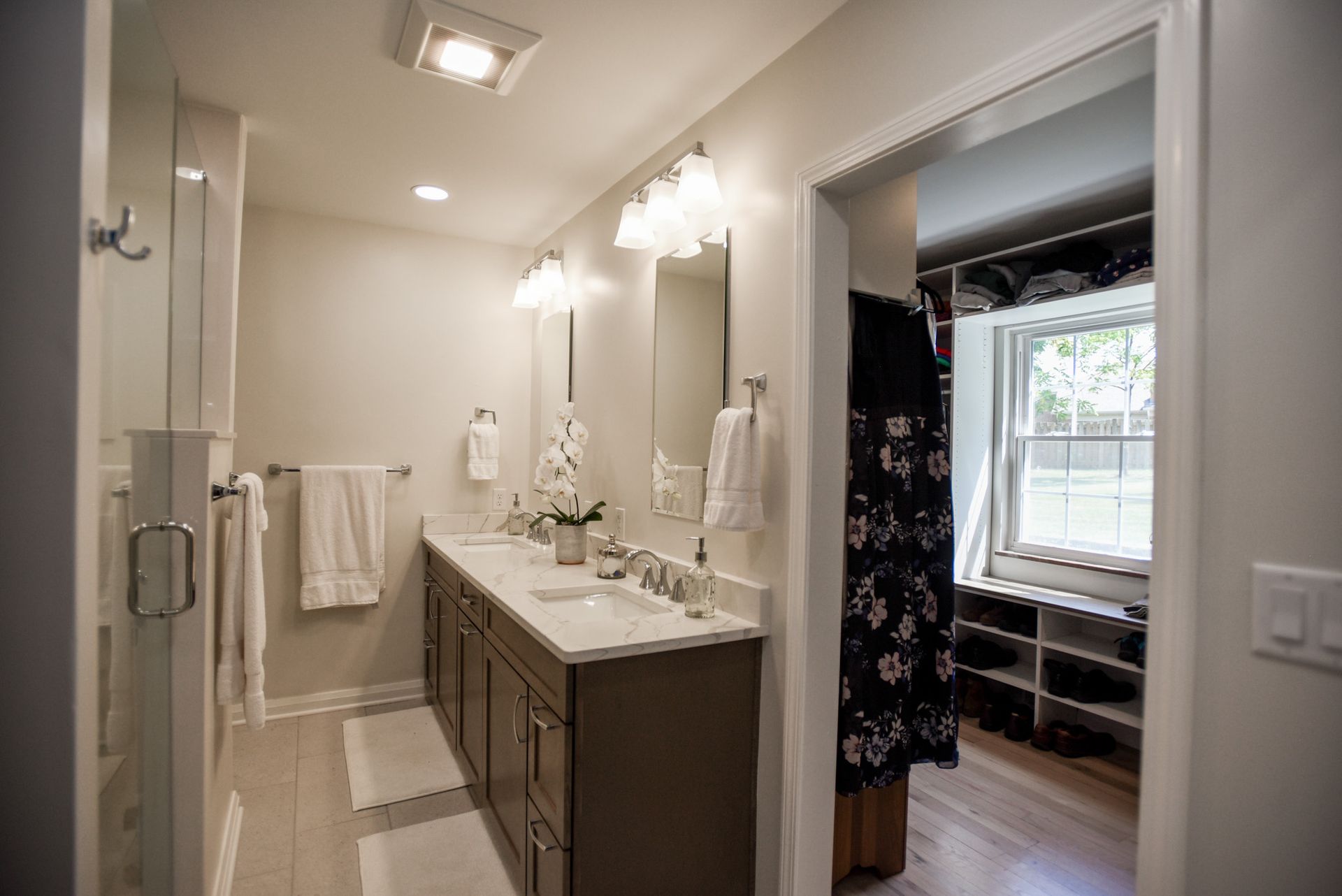 Bright bathroom with double vanity, mirrors, towel bar, and open doorway to a bedroom with a window.