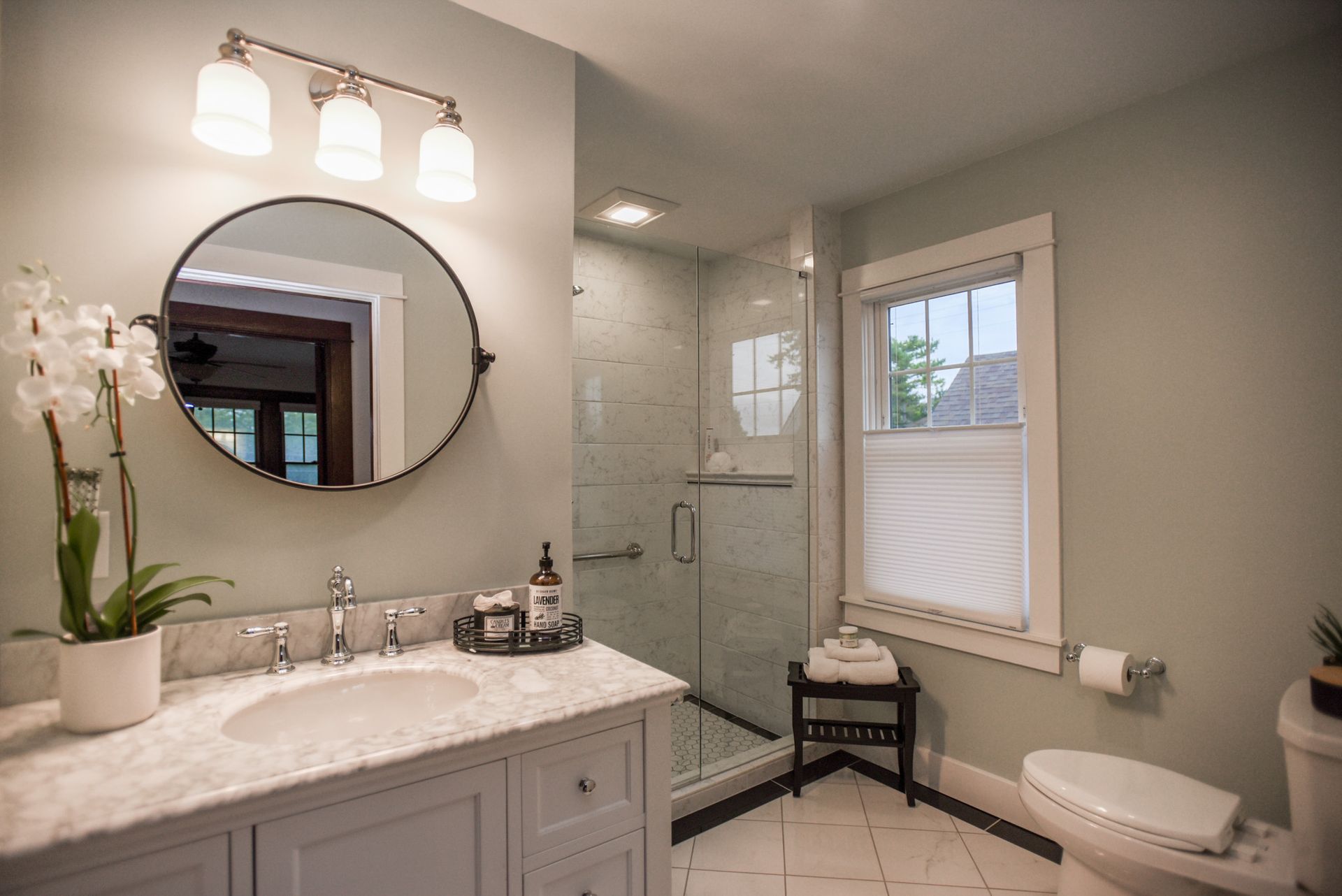 Bright bathroom with white vanity, round mirror, glass shower, window, and toilet under soft lighting.