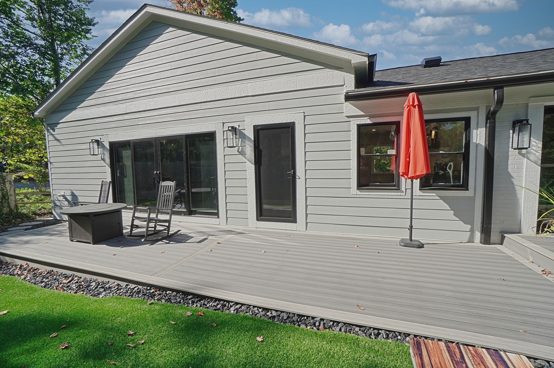 Deck with sliding glass doors, outdoor table and chairs, orange umbrella, and green lawn beside a house.