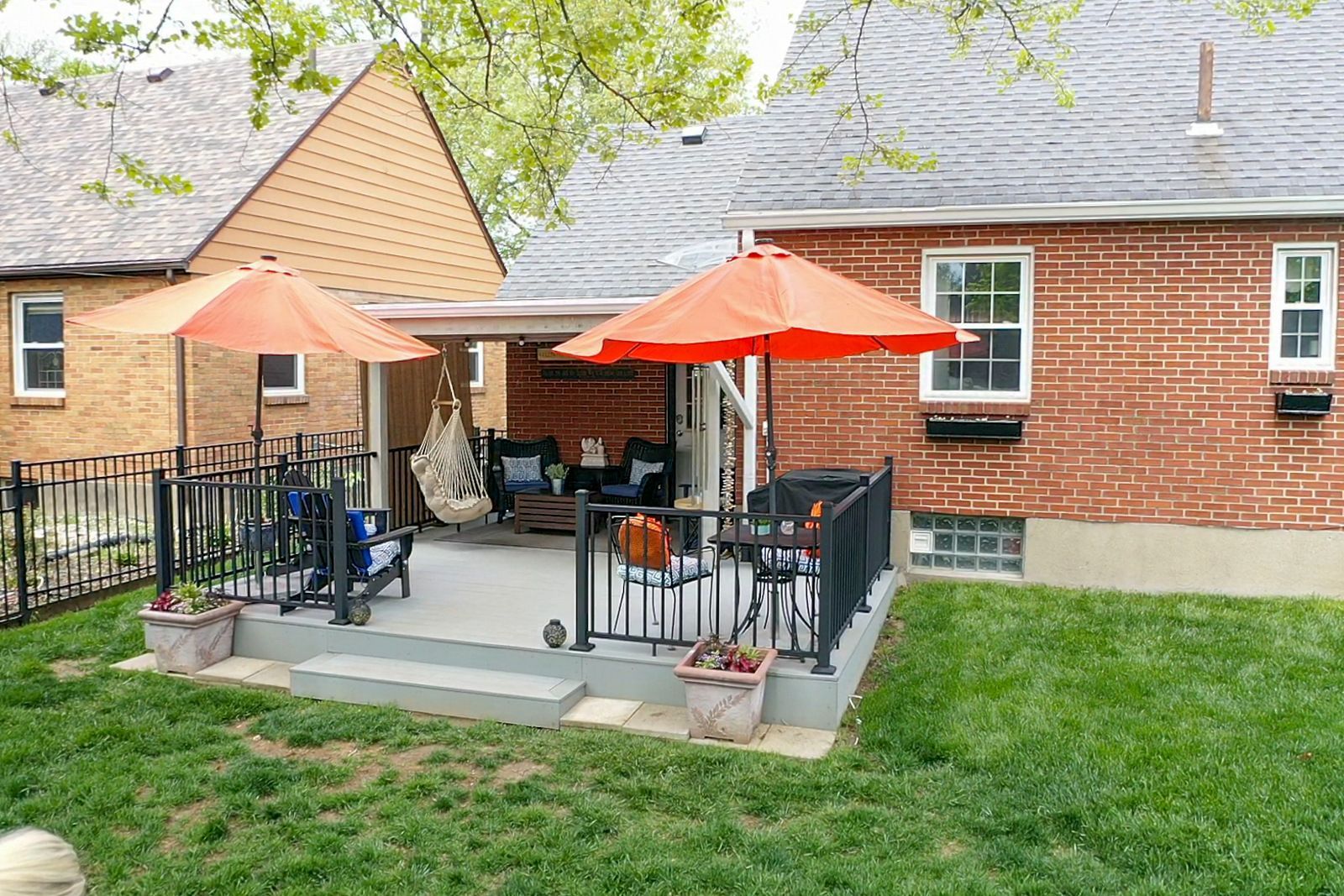 Backyard patio with tables, orange umbrellas, and black chairs beside a brick house.