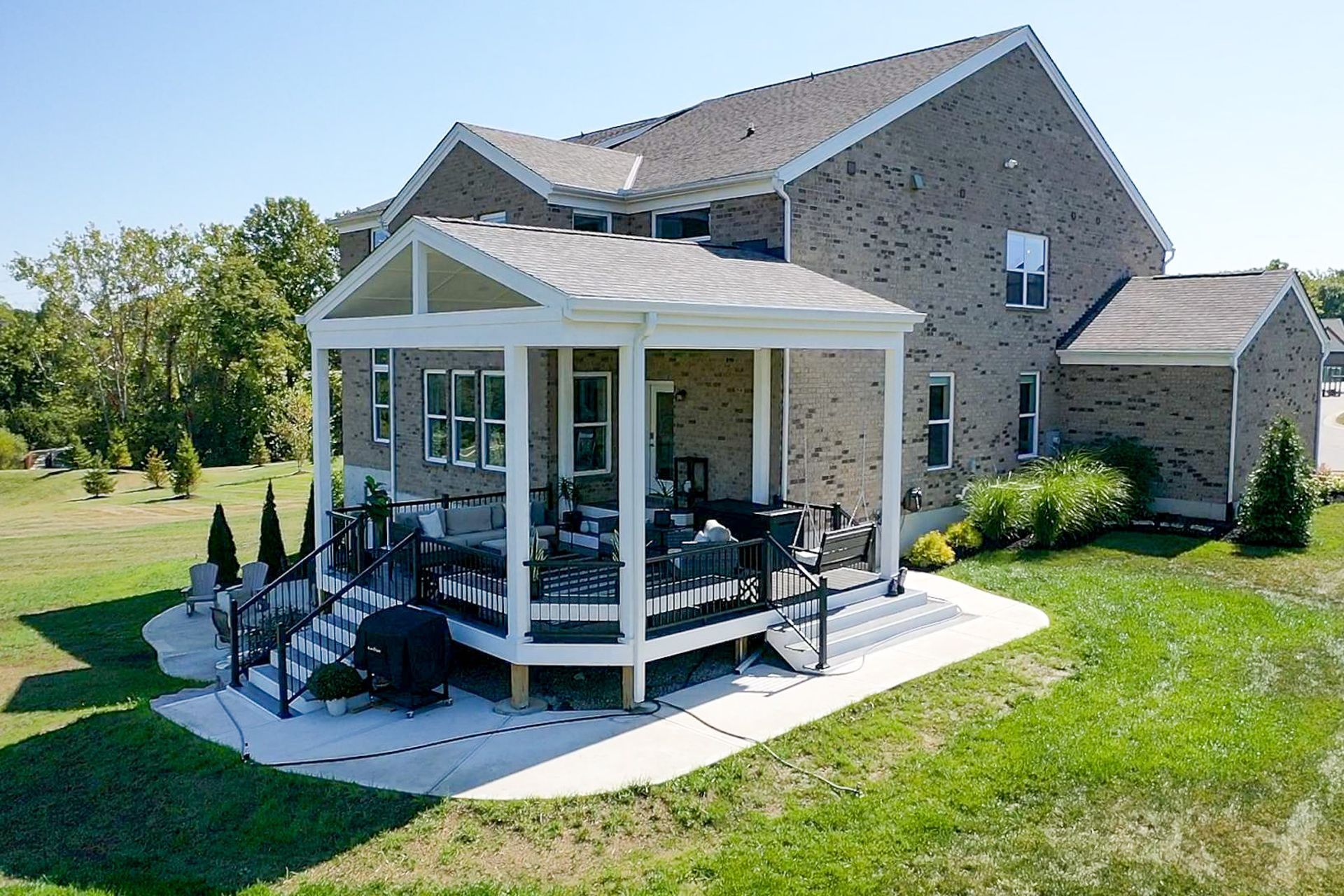Covered backyard patio with outdoor seating beside a brick house and green lawn.