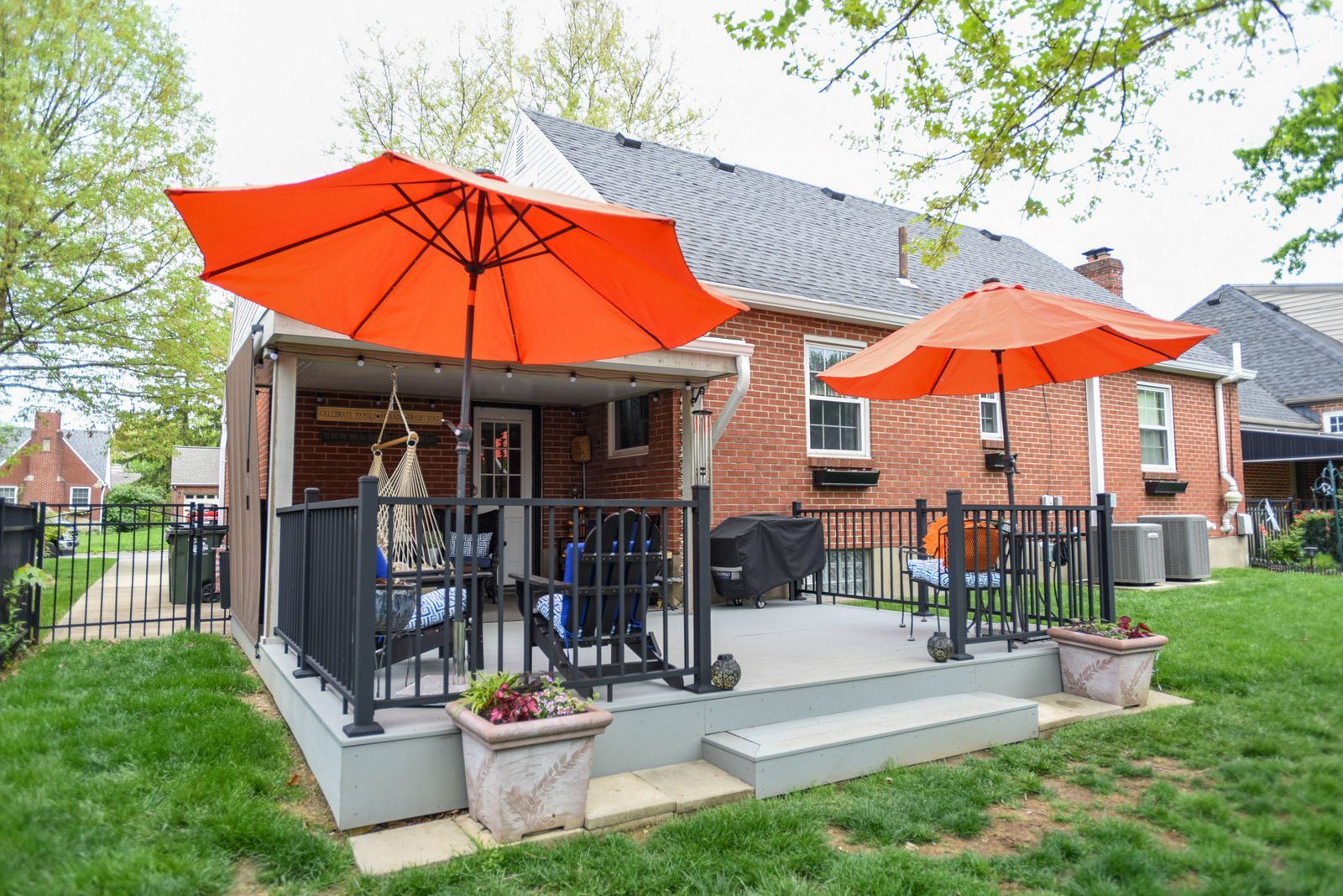 Backyard deck with orange umbrellas, black railing, and outdoor seating beside a brick house.