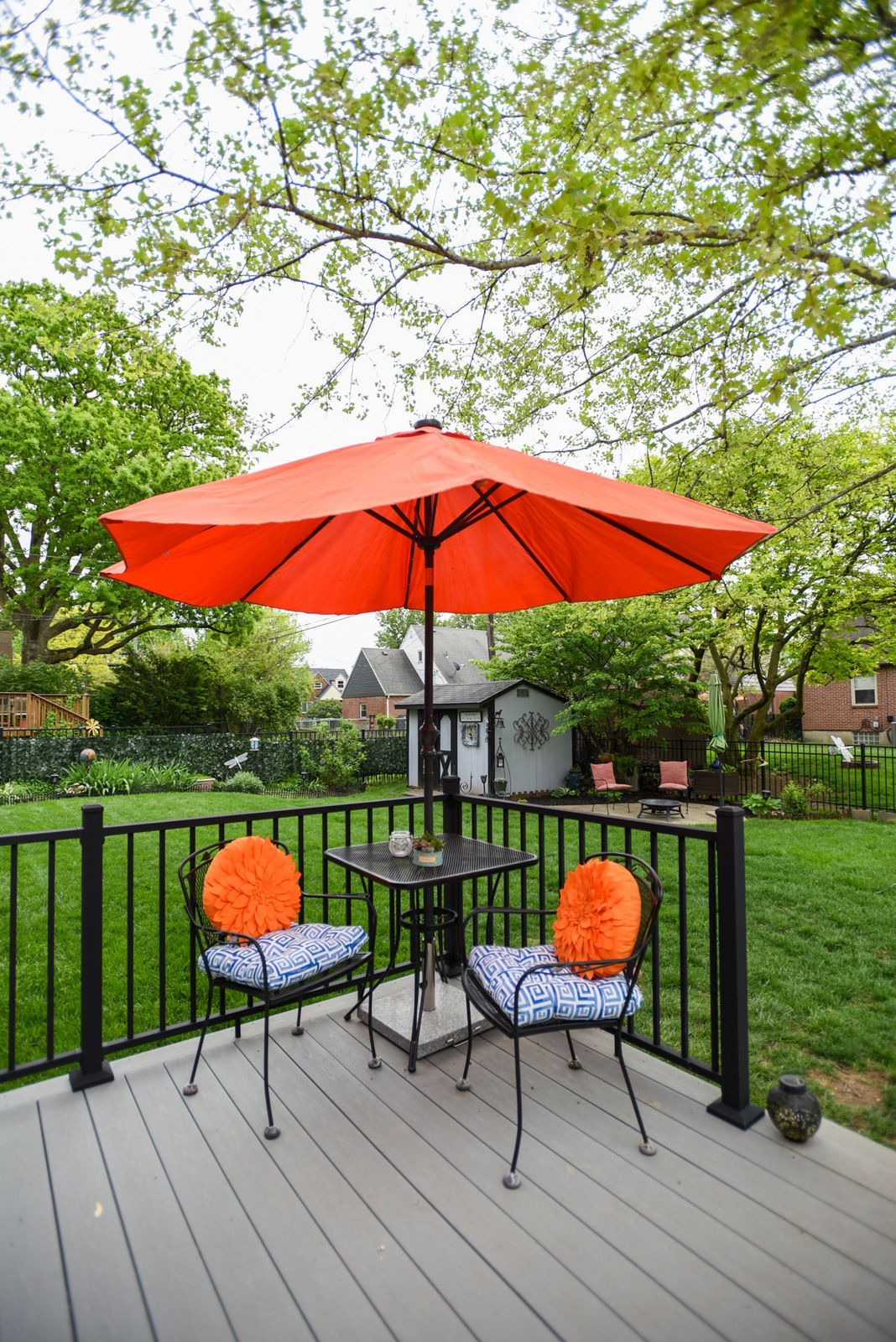 Patio deck with orange umbrella, two chairs, and small table overlooking a green backyard.