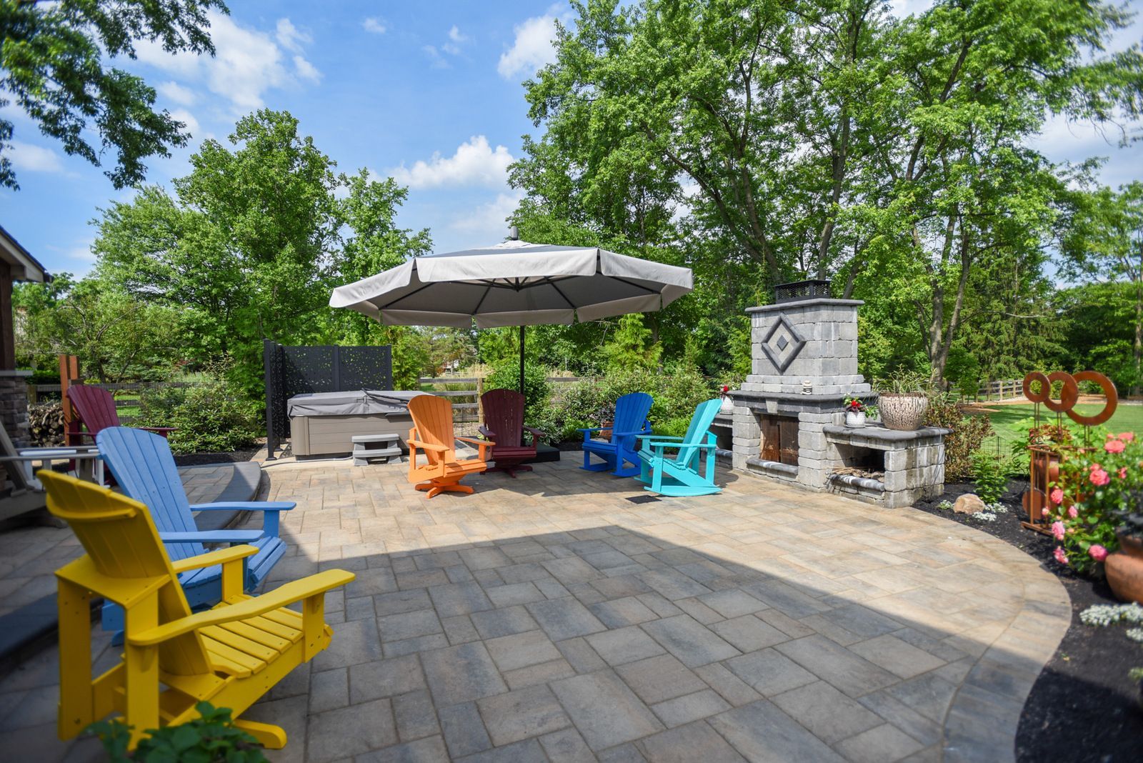 Colorful chairs around a patio fire pit and grill under an umbrella in a landscaped backyard.