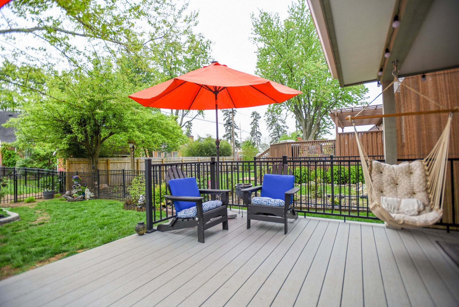 Backyard deck with two blue chairs, red umbrella, and hanging hammock beside a green yard.