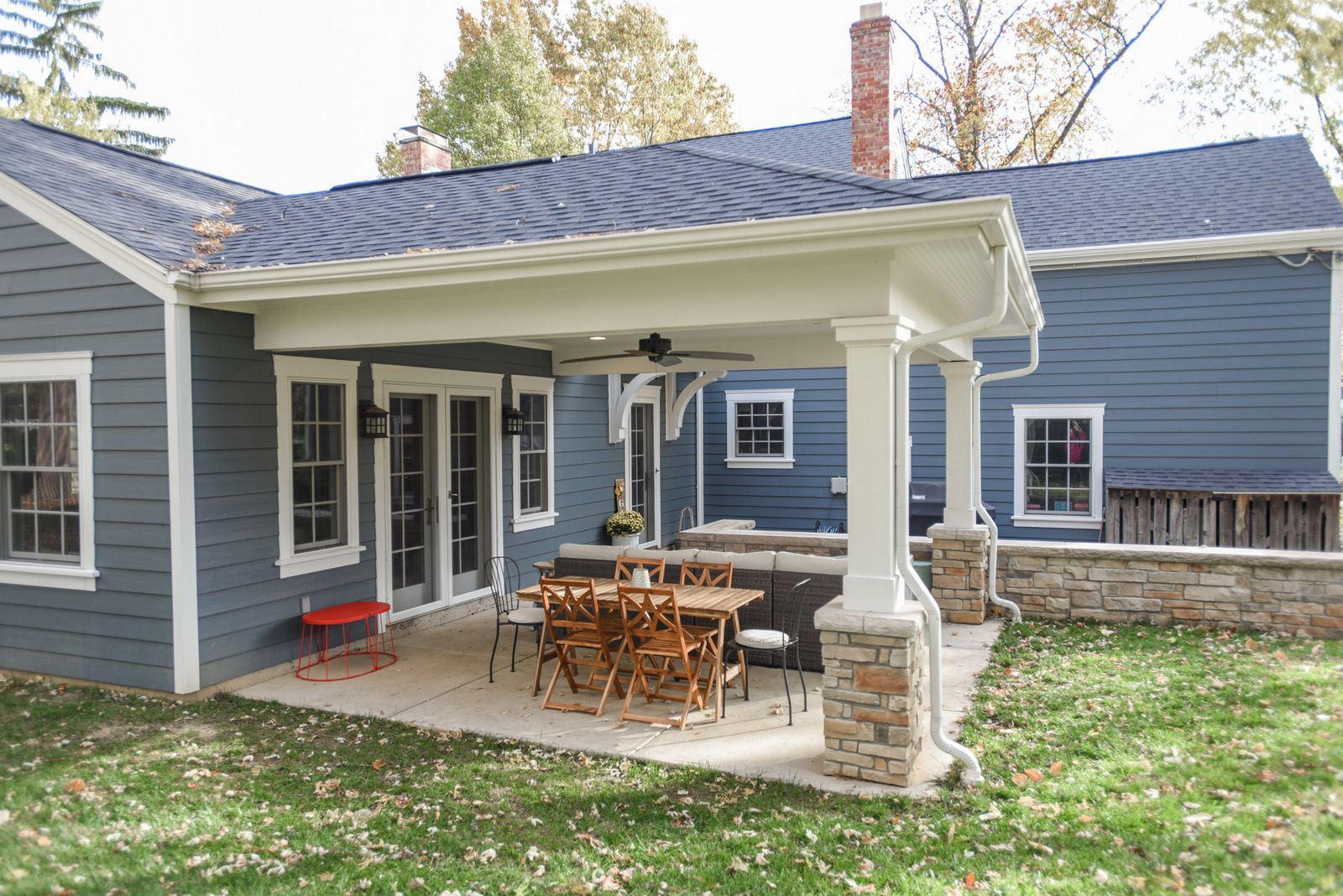 Back patio with covered porch, outdoor dining table, and blue house exterior amid autumn leaves.