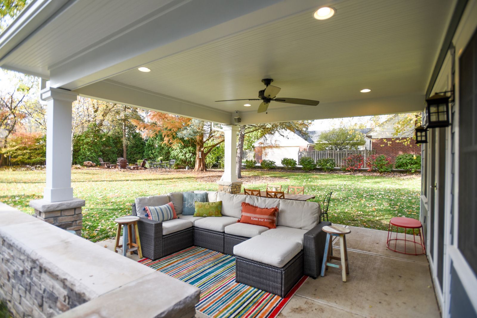 Covered patio with gray sectional sofa, colorful rug, and ceiling fan overlooking a grassy backyard.