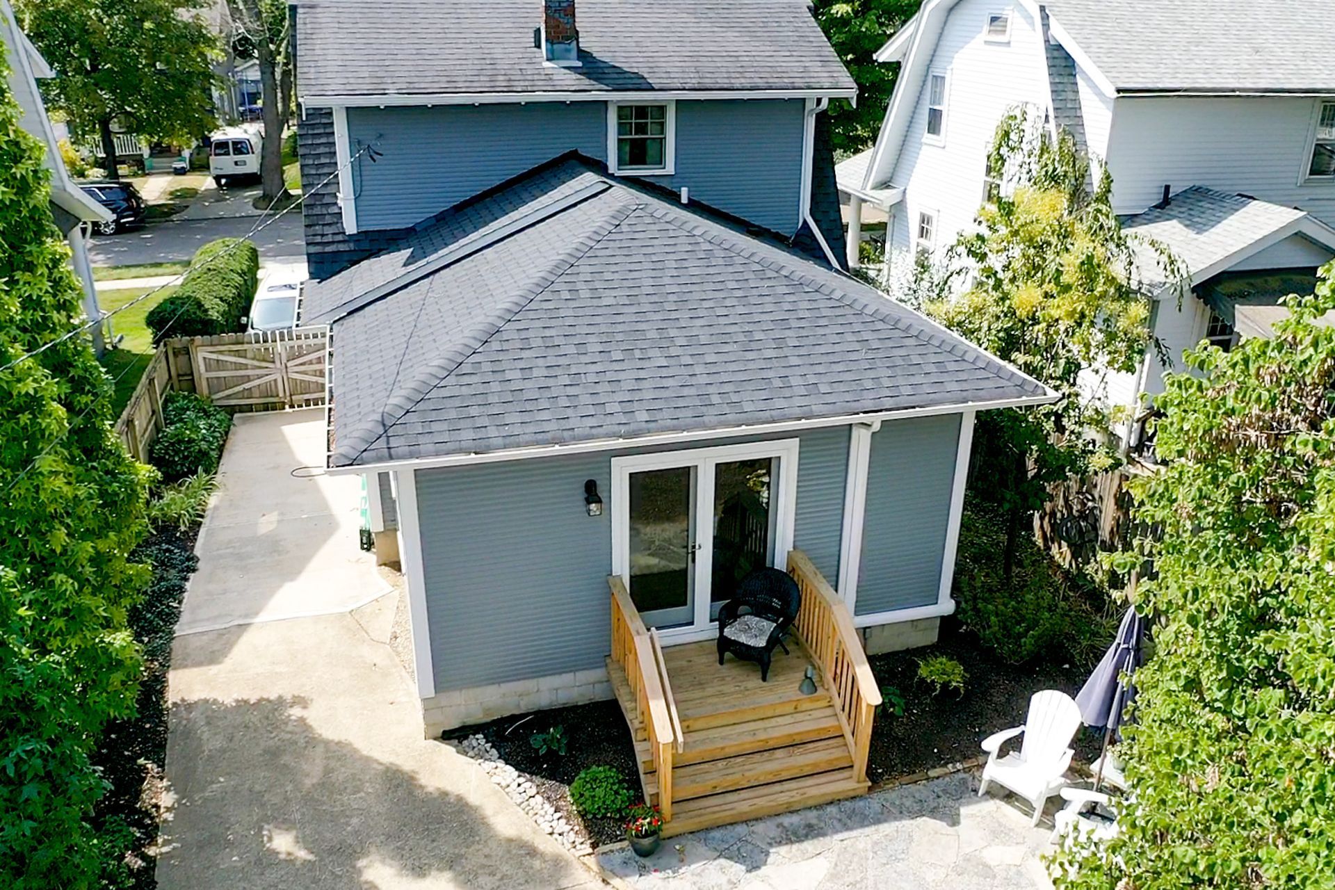 Aerial view of a small gray house with a wooden front deck and walkway, surrounded by trees and neighboring homes.
