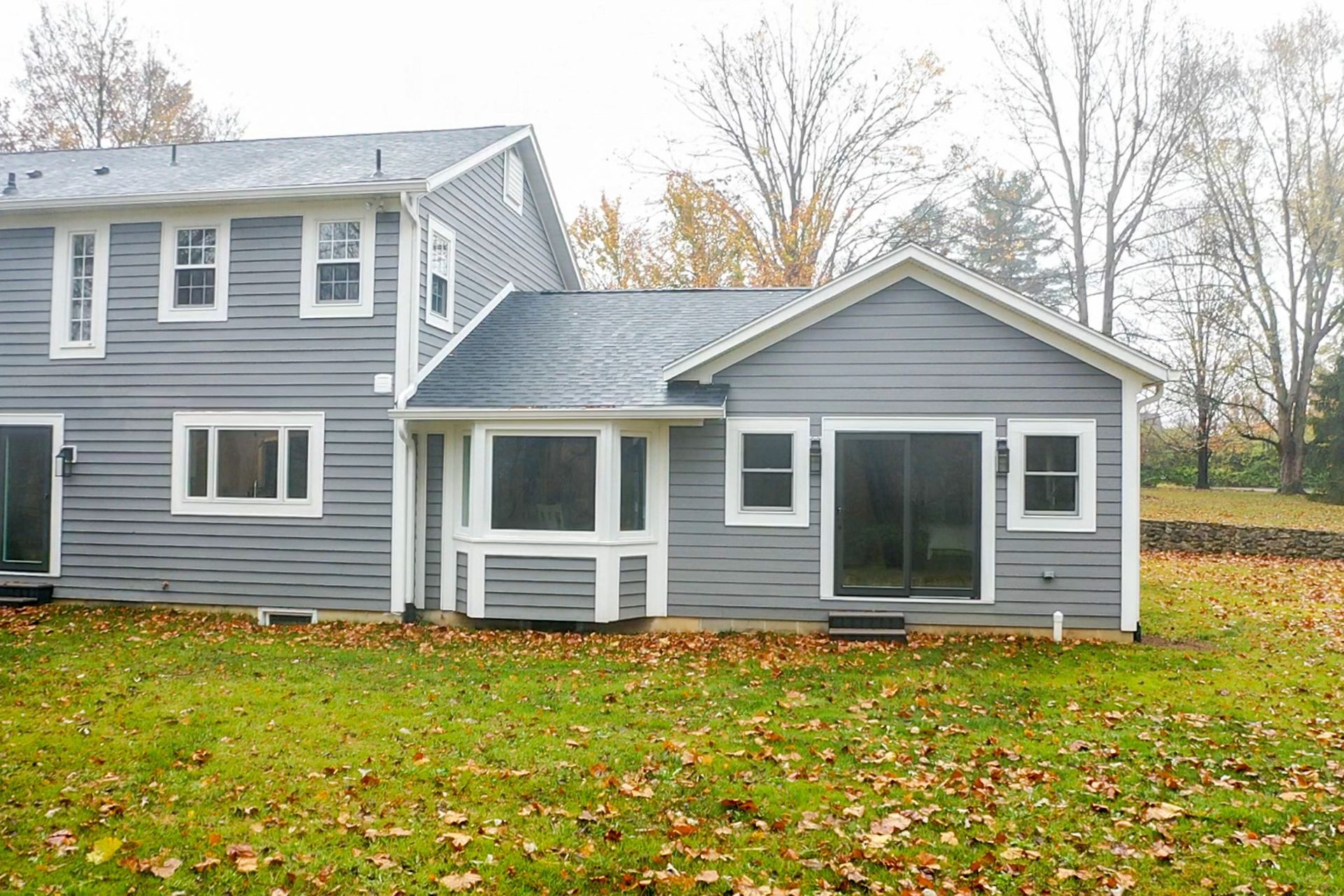 Gray two-story house with white trim and a small sunroom, set in a grassy yard with autumn trees.